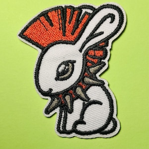 May include: An embroidered patch featuring a white rabbit character with a bright orange mohawk hairstyle. The rabbit has a black spiked collar around its neck and black outlines. The design is rendered in white, orange, black, and grey thread.