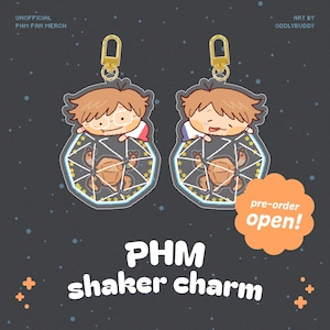 May include: Two clear acrylic shaker charms with gold-coloured clasps. Each charm features a cartoon character with brown hair and glasses, inside a geometric shape with a turtle design. Text on the image reads "PHM shaker charm" and "pre-order open!"