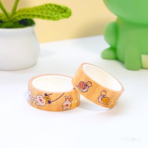 Op de afbeelding: Twee rollen decoratieve washi-tape. De tape is lichtbruin met cartoon illustraties van slakken en andere karakters. De tape ligt op een wit oppervlak met een groen kikker-vormig object op de achtergrond.