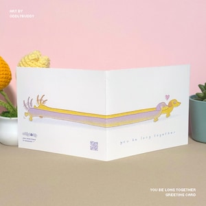 Peut inclure: Carte de vœux blanche avec une illustration de teckel de dessin animé. Le teckel est jaune et violet, avec le texte "you be long together". La carte est ouverte, avec l'art d'Oddlybuddy.