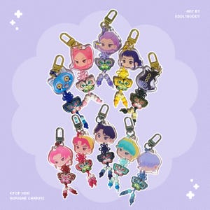 May include: A collection of nine acrylic charms featuring chibi-style character heads attached to colorful knotted cord tassels with butterfly-shaped accents and gold-toned lobster clasps. The text reads KPOP MINI NORIGAE CHARMS and ART BY ODDLYBUDDY.