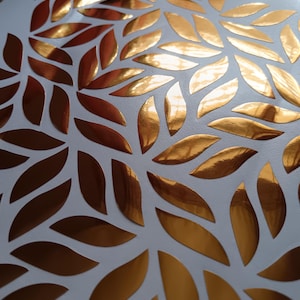 Può includere: Primo piano di una superficie bianca adornata da numerosi ritagli dorati a forma di foglia. Le foglie metalliche sono disposte in un motivo ripetuto, creando un design decorativo e accattivante. Il colore oro crea un contrasto sorprendente con lo sfondo bianco.