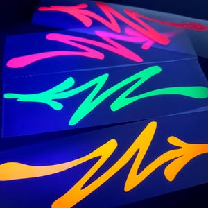 Könnte beinhalten: Drei rechteckige Aufkleber mit neonfarbenen Pfeilmotiven. Der oberste Aufkleber ist neonpink, der mittlere neongrün und der untere neonorange. Jeder Aufkleber zeigt eine einzelne, stilisierte, gezackte Pfeilgrafik auf dunklem Grund.