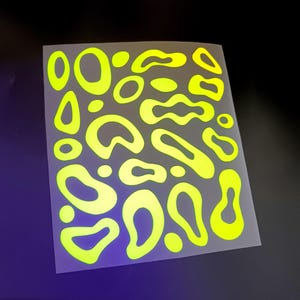 Può includere: Un foglio rettangolare con un motivo ripetitivo di forme organiche giallo neon simili ad amebe e piccoli cerchi pieni su uno sfondo grigio chiaro traslucido. Il design è composto da varie forme irregolari, fluide e arrotondate.