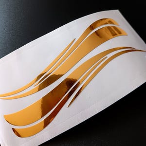 Op de afbeelding: Gouden metallic vinyl stickers met een vloeiend, golfachtig ontwerp. De stickers zitten op een witte achtergrond met een glanzende afwerking. Het ontwerp heeft meerdere gebogen lijnen, waardoor een dynamisch en opvallend beeld ontstaat.