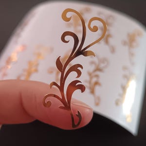 Könnte beinhalten: Nahaufnahme eines goldfarbenen, dekorativen Blumenaufklebers, der zwischen den Fingern gehalten wird. Der Aufkleber hat ein wirbelndes, verziertes Design mit Blättern und Locken. Eine Rolle ähnlicher Aufkleber ist im Hintergrund zu sehen, mit weißer Oberfläche und goldenen Blumenmustern.