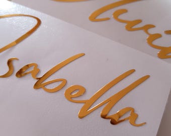 May include: Close-up of two white cards with gold foil lettering. The top card reads "Olivia" in cursive script. The bottom card reads "Isabella" in cursive script. The gold lettering is reflective and stands out against the white background.