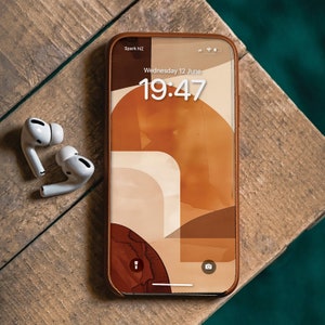Può includere: Uno smartphone con una custodia in pelle marrone mostra un design astratto dai toni caldi. Lo schermo mostra l'ora 19:47 di mercoledì 12 giugno. Auricolari wireless bianchi sono appoggiati accanto al telefono su una superficie di legno.