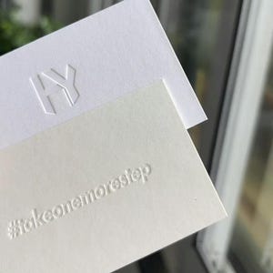 Op de afbeelding: Twee witte rechthoekige kaarten met reli&euml;fletters. De bovenste kaart bevat de letters HY in een geometrisch lettertype. De onderste kaart toont de hashtag #takeonemorestep in een strak, schreefloos lettertype.