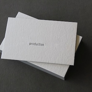 Op de afbeelding: Een stapel rechthoekige witte visitekaartjes met een getextureerde, zware papierafwerking. Het bovenste kaartje bevat het woord production gedrukt in een klein, zwart, schreefloos lettertype gecentreerd op het oppervlak.