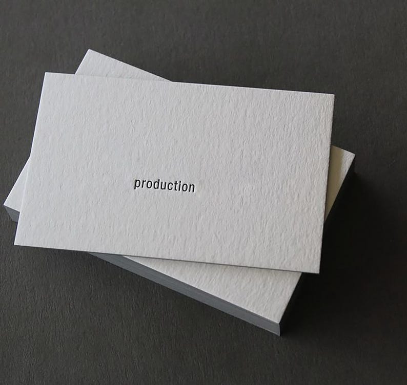 Op de afbeelding: Een stapel rechthoekige witte visitekaartjes met een getextureerde, zware papierafwerking. Het bovenste kaartje bevat het woord production gedrukt in een klein, zwart, schreefloos lettertype gecentreerd op het oppervlak.