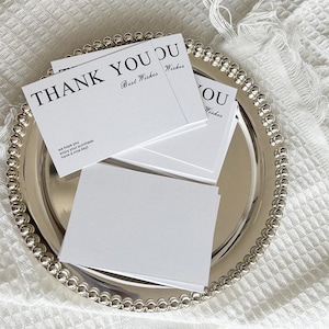 May include: A collection of white thank you cards, each bearing the words "THANK YOU" and "Best Wishes" in black. The cards rest on a silver tray with a beaded border. The cards also say "we hope you enjoy your purchase. Have a nice day!"