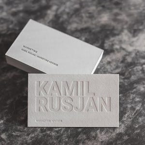 Könnte beinhalten: Weiße Visitenkarten mit dem geprägten Namen "KAMIL RUSJAN" auf der Vorderseite. Der Text "MARKETING ADVISOR" ist darunter gedruckt. Auf der Oberseite des Kartenstapels steht das Wort "MARKETING".