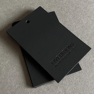 Könnte beinhalten: Zwei schwarze, rechteckige Anhänger mit abgerundeten Ecken, übereinander gestapelt. Jeder Anhänger hat ein kleines Loch oben und den geprägten Text "FIVEDITIONS WZ.FIVED1 DESIGN STUDIO". Die Anhänger liegen auf einer grauen Oberfläche.