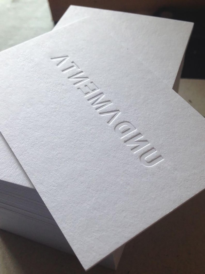 Op de afbeelding: Een stapel dikke, witte rechthoekige kaarten met een verdiepte afdruk van de tekst "UNDIVIDED" in hoofdletters. Het papier heeft een gestructureerde, matte afwerking en de letters zijn in het oppervlak van de bovenste kaart gedrukt.
