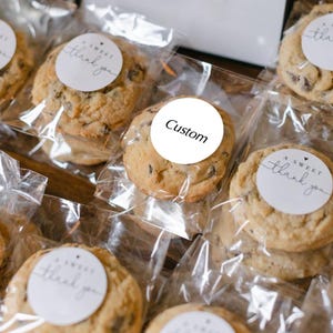 May include: Close-up of several chocolate chip cookies individually wrapped in clear cellophane. Each cookie has a white circular sticker with the words "A Sweet Thank You" or "Custom" printed on it. The cookies are a light brown colour.