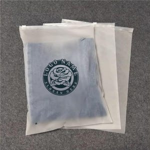 Puede incluir: Tres bolsas de ropa translúcidas con cremallera. Una bolsa contiene ropa doblada y presenta un logotipo circular con una rosa y el texto "LOGO NAME" y "SLOGAN HERE". Las otras dos bolsas están vacías y apiladas detrás de la primera.