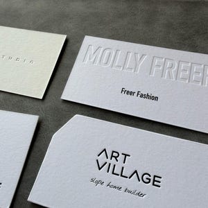 Op de afbeelding: Vier rechthoekige witte visitekaartjes met diverse ontwerpen. E&eacute;n kaart bevat de in reli&euml;f gedrukte tekst MOLLY FREER en Freer Fashion. Een andere toont ART VILLAGE en slope home builder. Een derde toont een gedeeltelijke pauwenveergrafiek.