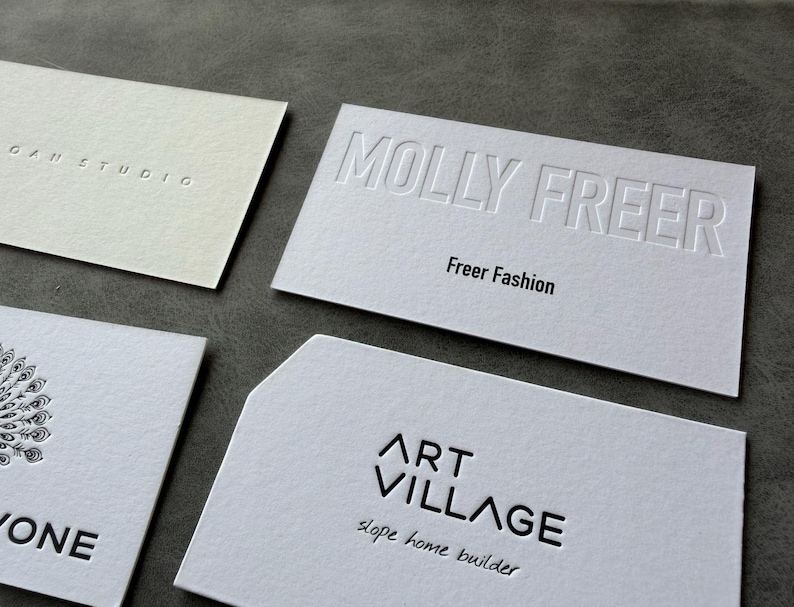 Op de afbeelding: Vier rechthoekige witte visitekaartjes met diverse ontwerpen. E&eacute;n kaart bevat de in reli&euml;f gedrukte tekst MOLLY FREER en Freer Fashion. Een andere toont ART VILLAGE en slope home builder. Een derde toont een gedeeltelijke pauwenveergrafiek.