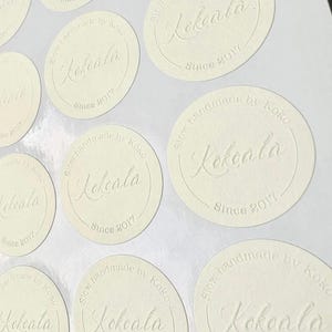 May include: Cream-coloured, circular stickers with embossed text. The stickers display the word "Kokeala" in a decorative font, with the text "Slow handmade by Koko Since 2017" around it. Several stickers are arranged on a white surface.