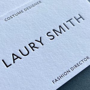 Op de afbeelding: Twee witte visitekaartjes met een gestructureerde papieren afwerking. De kaarten bevatten zwarte, verdiepte tekst met de tekst LAURY SMITH, FASHION DIRECTOR en COSTUME DESIGNER. De kaarten zijn licht versprongen op elkaar gestapeld.