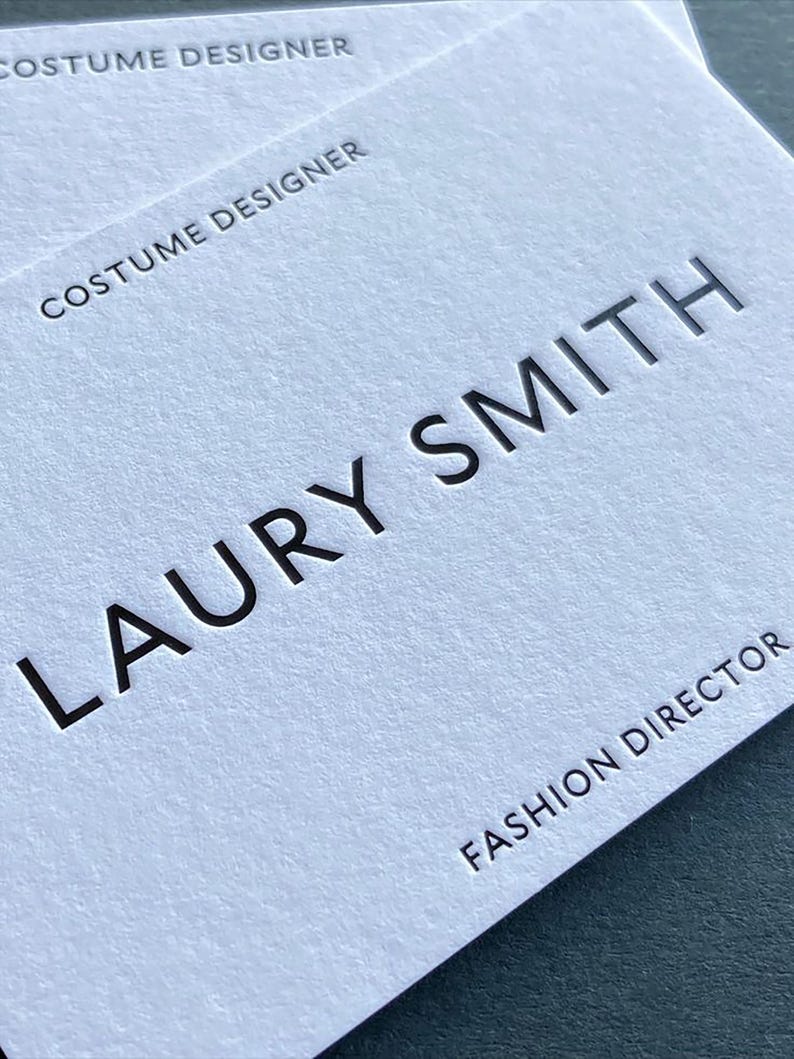 Op de afbeelding: Twee witte visitekaartjes met een gestructureerde papieren afwerking. De kaarten bevatten zwarte, verdiepte tekst met de tekst LAURY SMITH, FASHION DIRECTOR en COSTUME DESIGNER. De kaarten zijn licht versprongen op elkaar gestapeld.