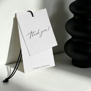 Pode incluir: Uma etiqueta de papel retangular branca com um cordão preto. A etiqueta apresenta o texto Thank you! escrito à mão com tinta preta no topo e o texto impresso ISHMAELCODESIGN em letras maiúsculas pretas na parte inferior central.