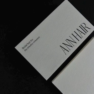 Op de afbeelding: Een rechthoekig wit visitekaartje met zwarte tekst. Het kaartje bevat de naam ANN HAIR in een groot schreeflettertype en de zin Bridal hair for the timeless romantic in een kleiner lettertype. Het kaartje ligt op een zwarte achtergrond.