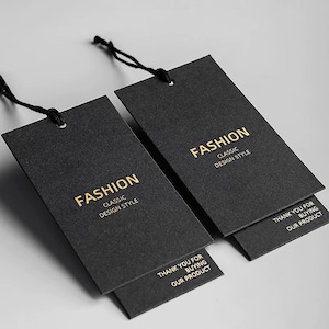 May include: Two black rectangular clothing tags with the word "FASHION" in gold lettering. The tags also read "CLASSIC DESIGN STYLE" and "THANK YOU FOR BUYING OUR PRODUCT". The tags are connected with black string.
