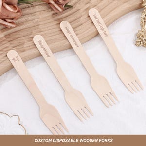 May include: Four wooden forks with different personalized messages: "Your text / logo here", "Happy anniversary", "Happy birthday", and "It's a girl". The forks are on a wooden surface.