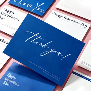 May include: A collection of greeting cards with white text on blue and white backgrounds. The cards feature phrases like "Thank you!", "Happy Valentine's Day", and "I love You". The cards are arranged on a pink surface.