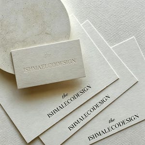 Könnte beinhalten: Eine Sammlung cremefarbener, rechteckiger Visitenkarten mit dem schwarzen Aufdruck "the ISHMAELCODESIGN", wobei eine Karte denselben Text in geprägter Form zeigt, alles angeordnet neben einer runden, strukturierten, cremefarbenen Steinscheibe.