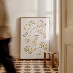Op de afbeelding: Een witte geframed poster met een gouden rand en een witte achtergrond. De poster toont een tekening van verschillende soorten brood in goud, met de woorden "EAT Bread" in zwart.
