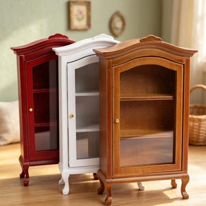 Pode incluir: Três vitrines em miniatura lado a lado, com acabamentos em vermelho, branco e madeira natural. Cada armário possui uma porta com painel de vidro, um pequeno puxador dourado, duas prateleiras internas e quatro pernas curvas.