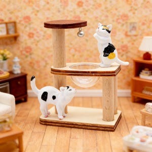 Könnte beinhalten: Miniatur-Katzenmöbel-Set mit zwei weißen und schwarzen Katzenfiguren. Das Set enthält einen Katzenbaum aus Holz mit brauner Oberseite, eine transparente Schüssel und eine hängende silberne Kugel.