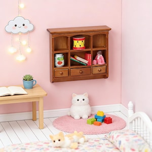 May include: A miniature wooden bookshelf with three drawers and open shelves, filled with tiny toys and books. A plush white cat sits on a pink rug, with colorful building blocks nearby. A small wooden table holds a blue cup and an open book.