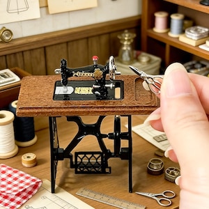 K&ouml;nnte beinhalten: Miniatur-N&auml;hmaschine mit einem Holztisch und schwarzem Metallgestell. Die Maschine ist schwarz mit goldenen Akzenten und einem roten Knopf. Zubeh&ouml;r umfasst Garnrollen, Scheren und ein Lineal, was auf ein Bastel- oder Hobbythema hindeutet.
