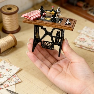 K&ouml;nnte beinhalten: Miniatur-N&auml;hmaschine in Schwarz mit goldenen Akzenten, ein rot-wei&szlig; kariertes Tuch und eine kleine Schere. Die N&auml;hmaschine steht auf einem Holztisch mit schwarzem Metallgestell. Garnrollen und florale Stoffreste sind ebenfalls zu sehen.