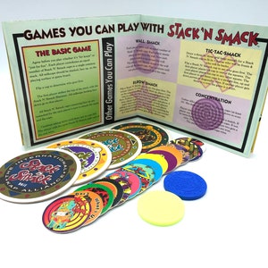 May include: A set of colorful game pieces with a variety of designs, including a green and yellow piece with the words "Stack 'N Smack" and a blue piece with the words "International Kap Alliance". The pieces are arranged on a white surface next to a book with instructions for playing different games with the pieces.