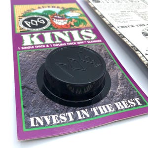 May include: A black plastic Kinis slammer with the POG logo, resting on a purple and green card. The card reads "KINIS" and "INVEST IN THE BEST".