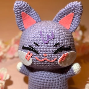 Può includere: Un giocattolo a forma di gatto di colore viola lavorato a uncinetto con orecchie rosa e un viso sorridente. Il gatto è seduto su un letto di fiori rosa.