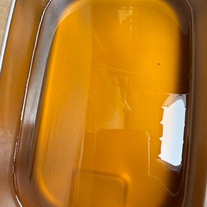 May include: A rectangular stainless steel container filled with a translucent amber-colored liquid. The metal basin features rounded corners and smooth, reflective walls that contain the golden-toned fluid, creating a clear surface reflection.