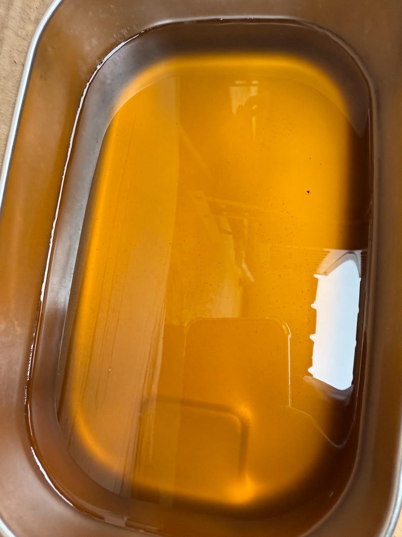 May include: A rectangular stainless steel container filled with a translucent amber-colored liquid. The metal basin features rounded corners and smooth, reflective walls that contain the golden-toned fluid, creating a clear surface reflection.