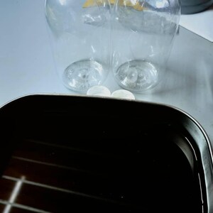 May include: Two clear, empty plastic bottles with rounded bases and narrow necks are positioned behind a rectangular, dark-colored metal tray filled with a dark liquid. Two small, white, circular caps are placed between the bottles and the tray.