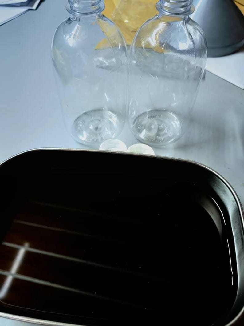 May include: Two clear, empty plastic bottles with rounded bases and narrow necks are positioned behind a rectangular, dark-colored metal tray filled with a dark liquid. Two small, white, circular caps are placed between the bottles and the tray.