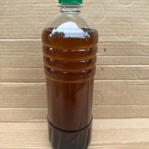May include: A clear plastic bottle filled with a dark amber liquid, featuring a ribbed cylindrical body and a solid green screw-top cap. The container is transparent, allowing the deep brown color of the liquid inside to be clearly visible.