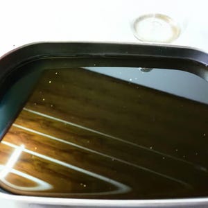 May include: A rectangular electric griddle with rounded corners and a black non-stick surface. The cooking area is filled with a dark, reflective liquid, showing a clear reflection of the surrounding environment on its smooth, glossy surface.