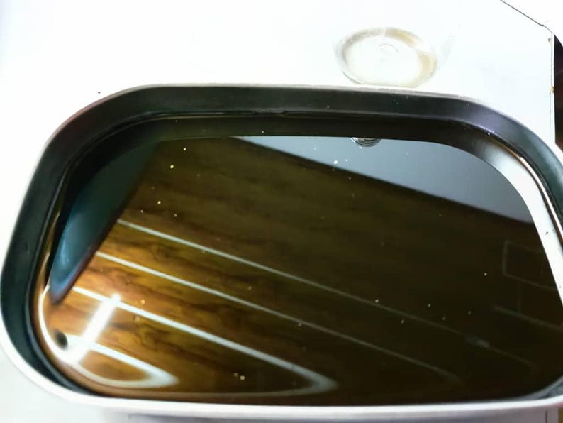 May include: A rectangular electric griddle with rounded corners and a black non-stick surface. The cooking area is filled with a dark, reflective liquid, showing a clear reflection of the surrounding environment on its smooth, glossy surface.