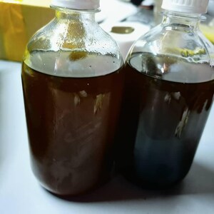May include: Two clear glass bottles with white screw-top caps, each filled with a dark brown liquid. The bottles have a rounded shoulder and a cylindrical body, containing a translucent, amber-toned fluid that appears consistent in both containers.