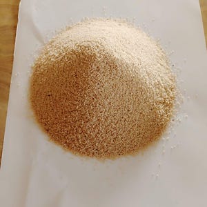 May include: A pile of light brown, granular material, possibly a spice or ingredient, sits on a white sheet of paper. The material is finely textured and appears to be a dry, powdery substance. The paper is on a light-colored wooden surface.